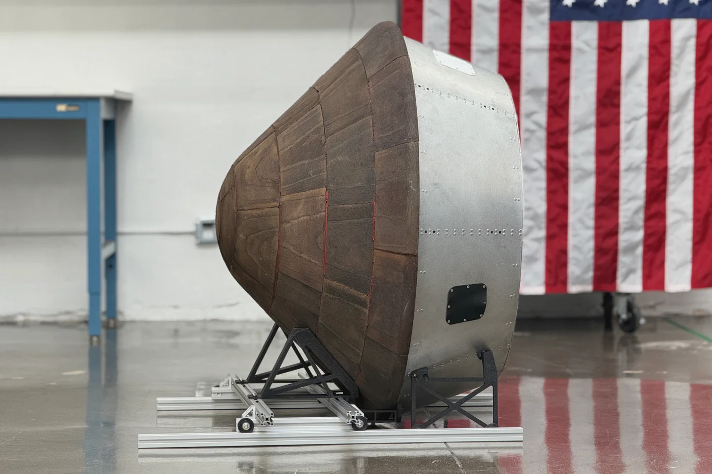 The full-scale Spark-1 prototype on an aluminum cradle on a polished shop floor: the segmented tan ablative heat shield forward, the riveted bare-aluminum backshell with a black access hatch, and a large United States flag hanging behind